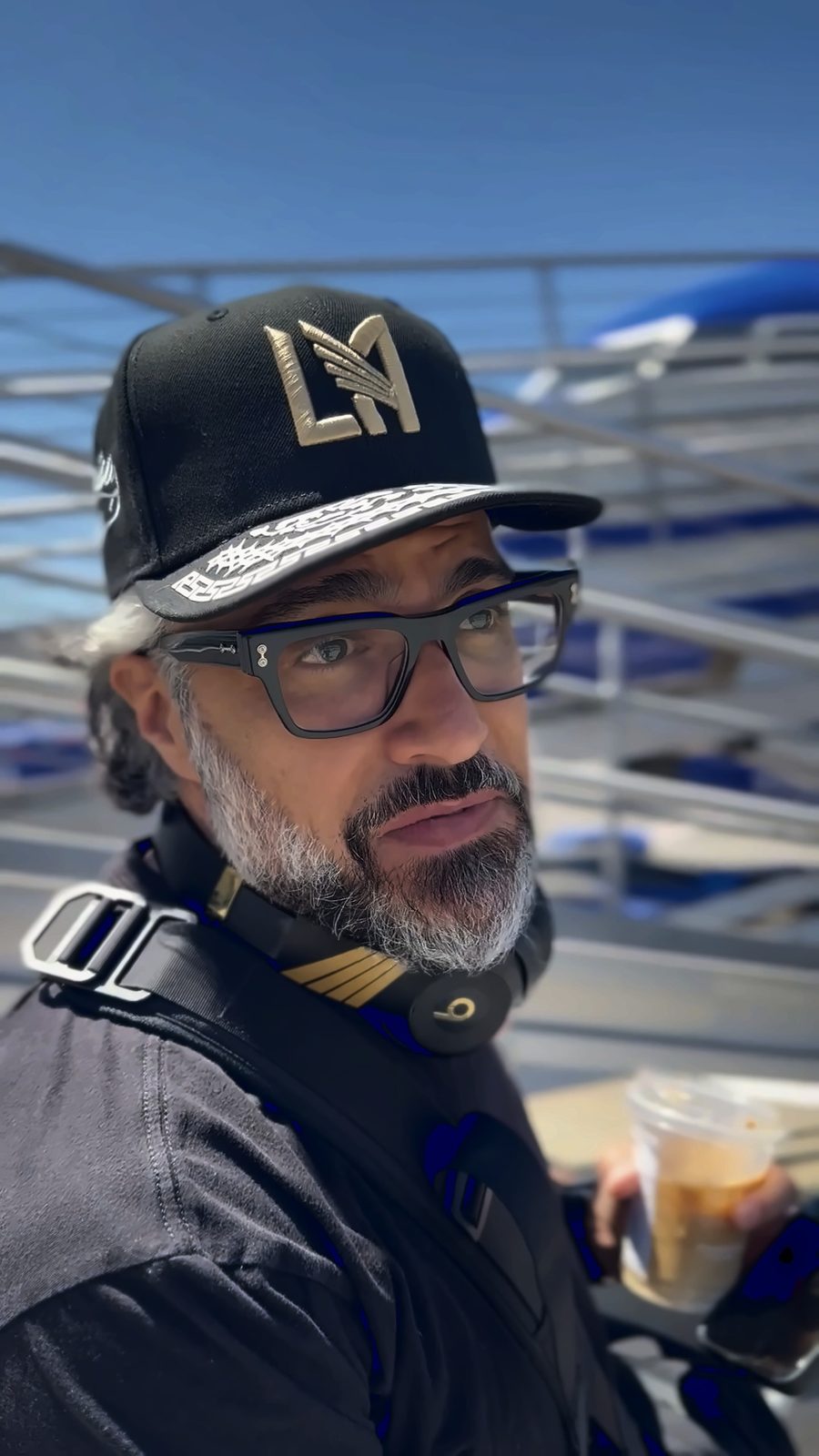 Jaime Camil in an LAFC cap and glasses, holding an iced coffee on the tarmac at Burbank airport